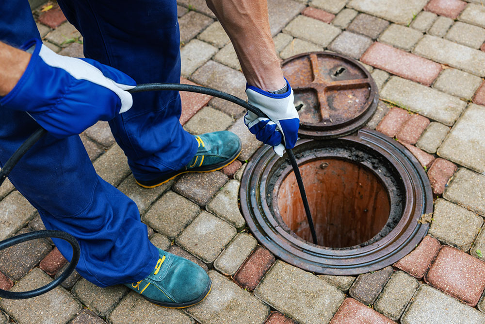Worker cleaning sewer drain with hose, wearing gloves and boots on paved surface.