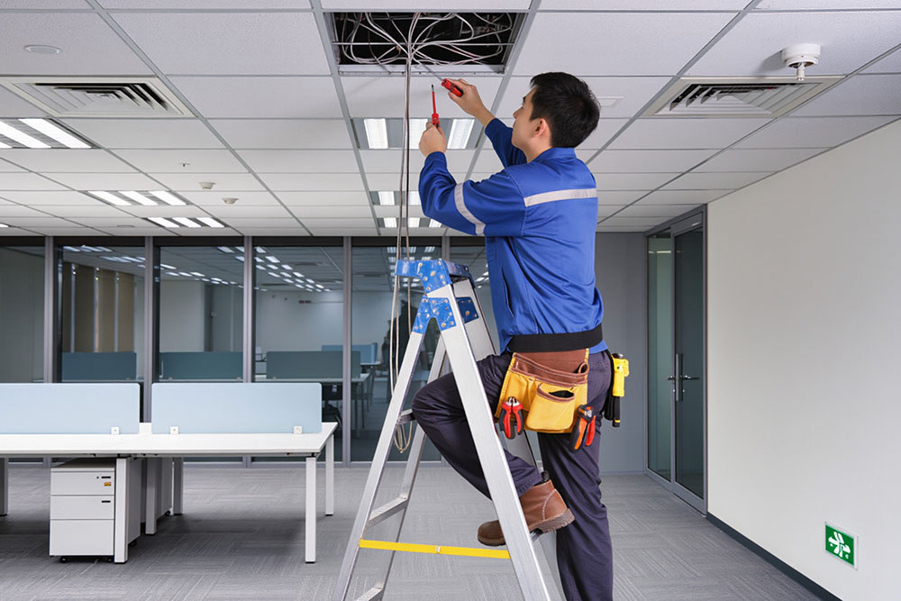 Technician on ladder repairing office ceiling wires, showcasing electrical maintenance.