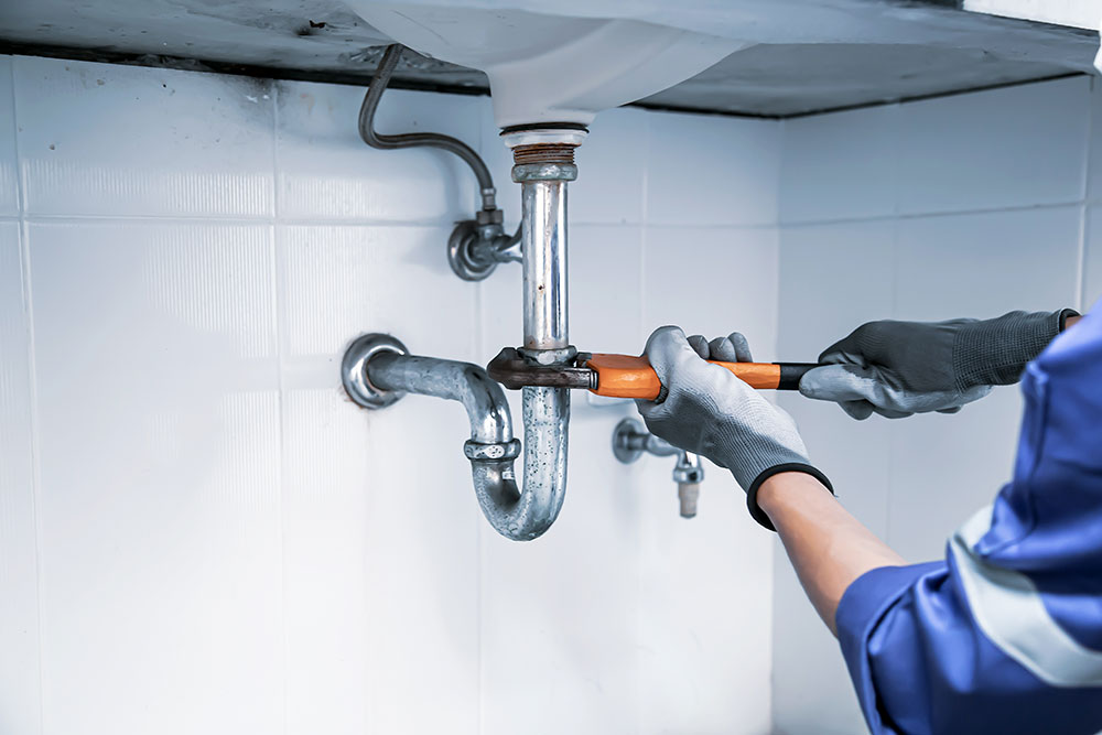 Plumber fixing a sink pipe with a wrench, wearing gloves for safety in a tiled bathroom setting.