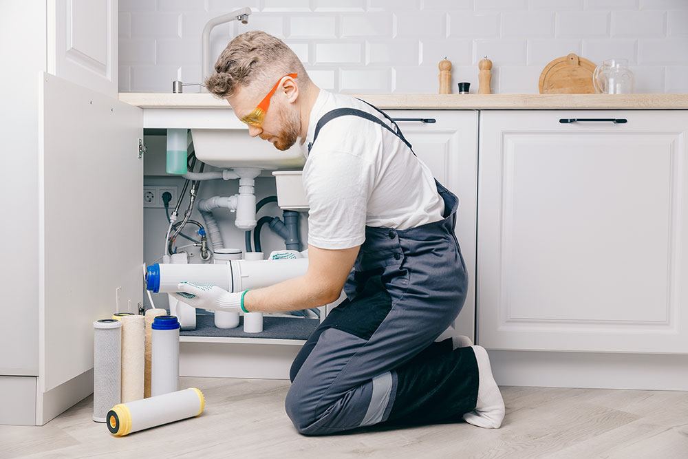 Technician installing water filter under kitchen sink, wearing safety glasses and overalls.