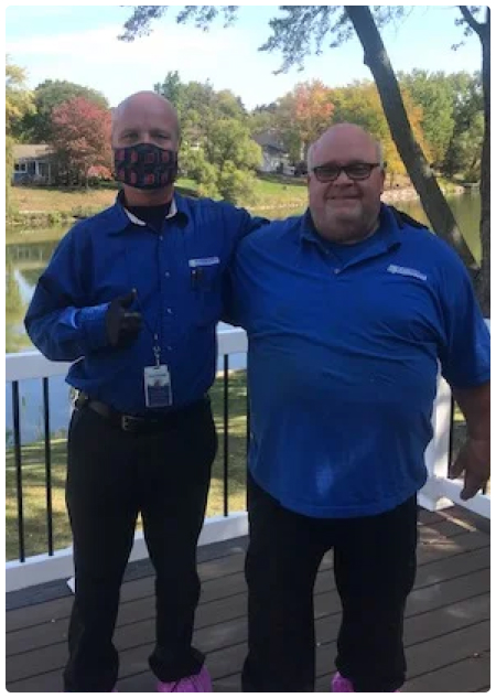 Two men standing together on a deck with a scenic backdrop, both wearing blue shirts and smiling at the camera.