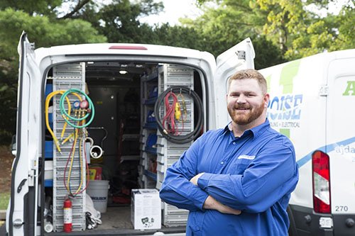 Smiling electrician in blue shirt standing by van stocked with equipment and tools, ready for service.