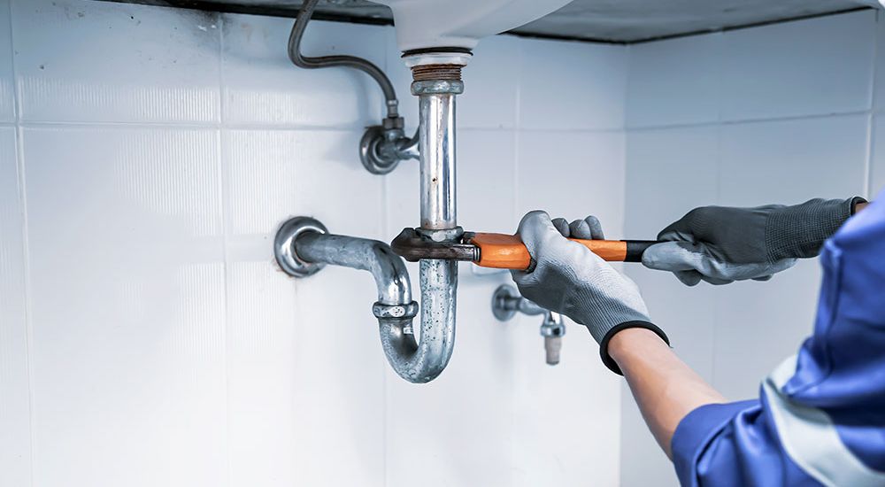Plumber fixing a sink pipe with a wrench, wearing gloves for safety in a tiled bathroom setting.