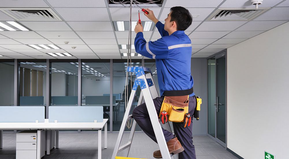 Technician on ladder repairing office ceiling wires, showcasing electrical maintenance.