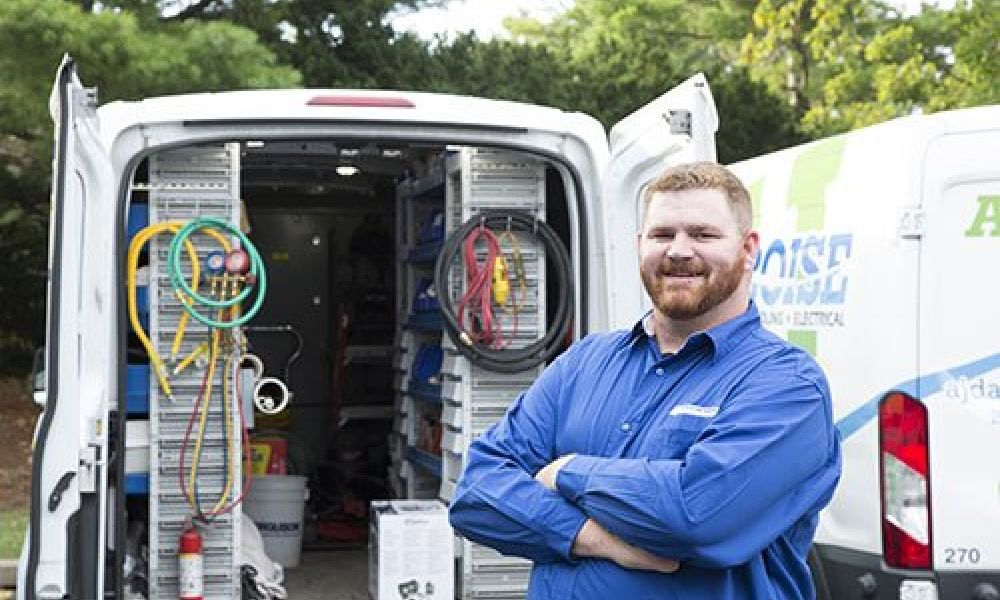 Smiling electrician in blue shirt standing by van stocked with equipment and tools, ready for service.