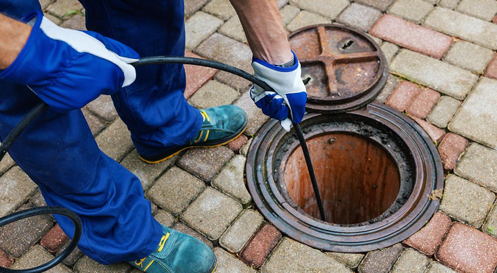 Worker cleaning sewer drain with hose, wearing gloves and boots on paved surface.