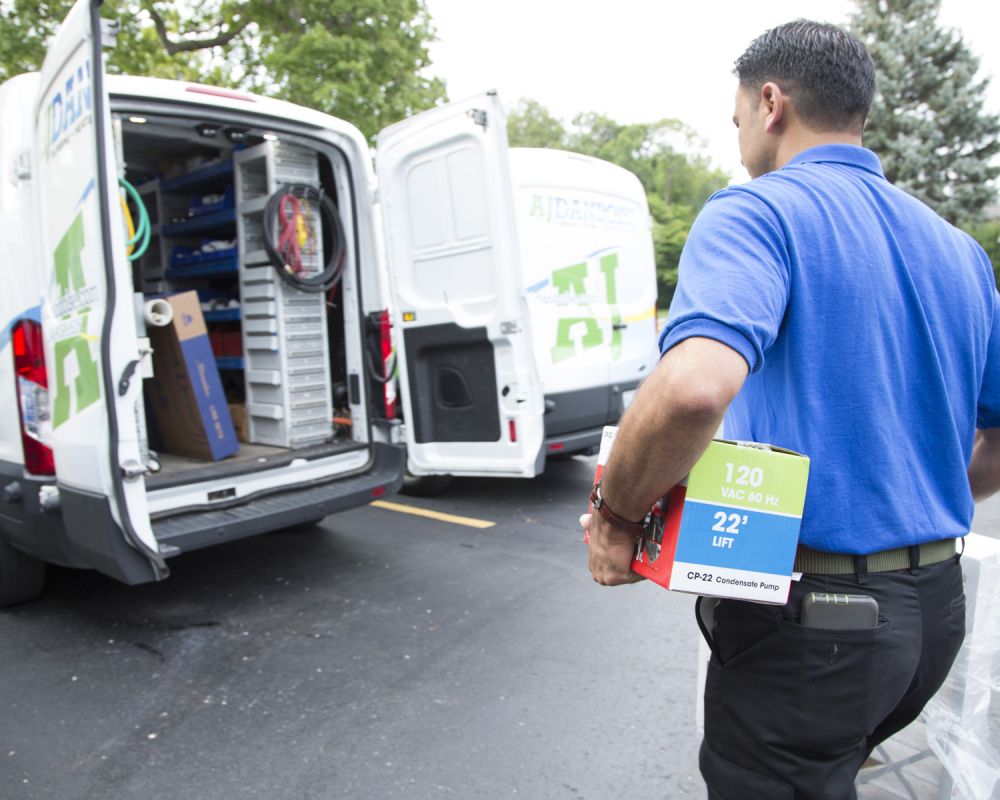 A worker in a blue shirt carries a boxed CP-22 condensate pump toward a white service van with its rear doors open and tools visible inside.