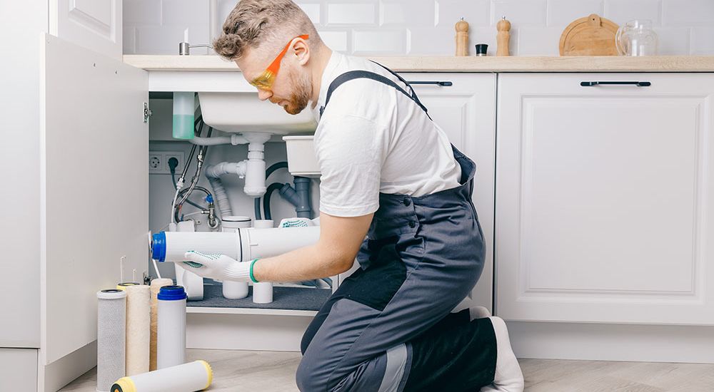 Technician installing water filter under kitchen sink, wearing safety glasses and overalls.