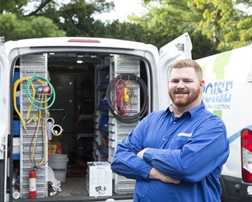 Smiling electrician in blue shirt standing by van stocked with equipment and tools, ready for service.