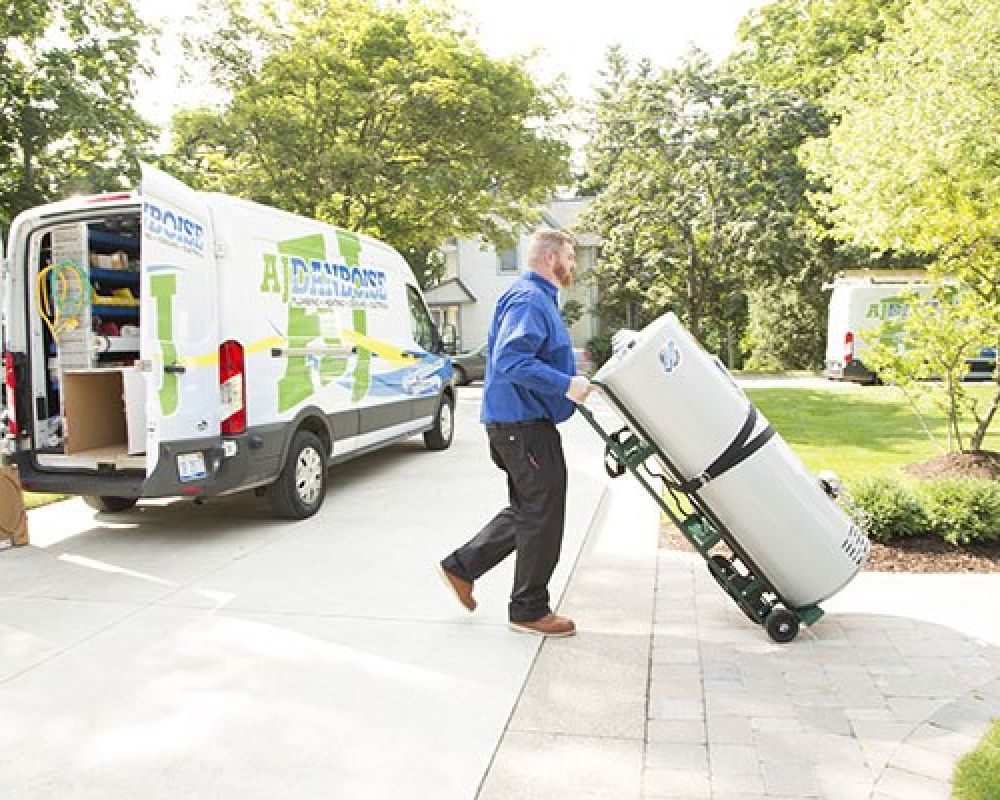 Technician delivering water heater using dolly to residential home from branded service van in sunny neighborhood.